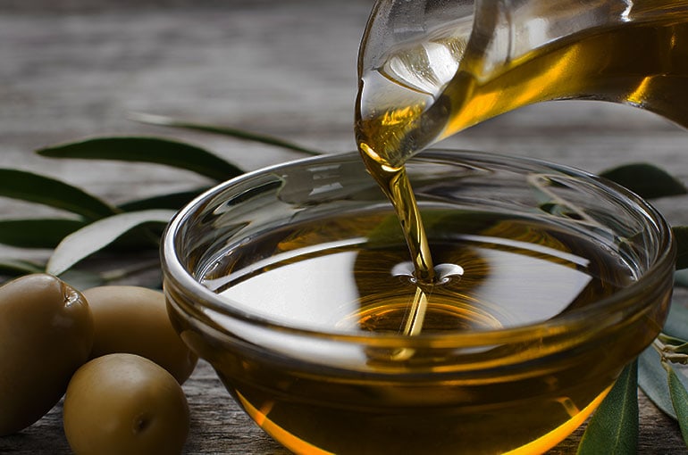 Oil poured from a glass decanter to a glass bowl next to olives.